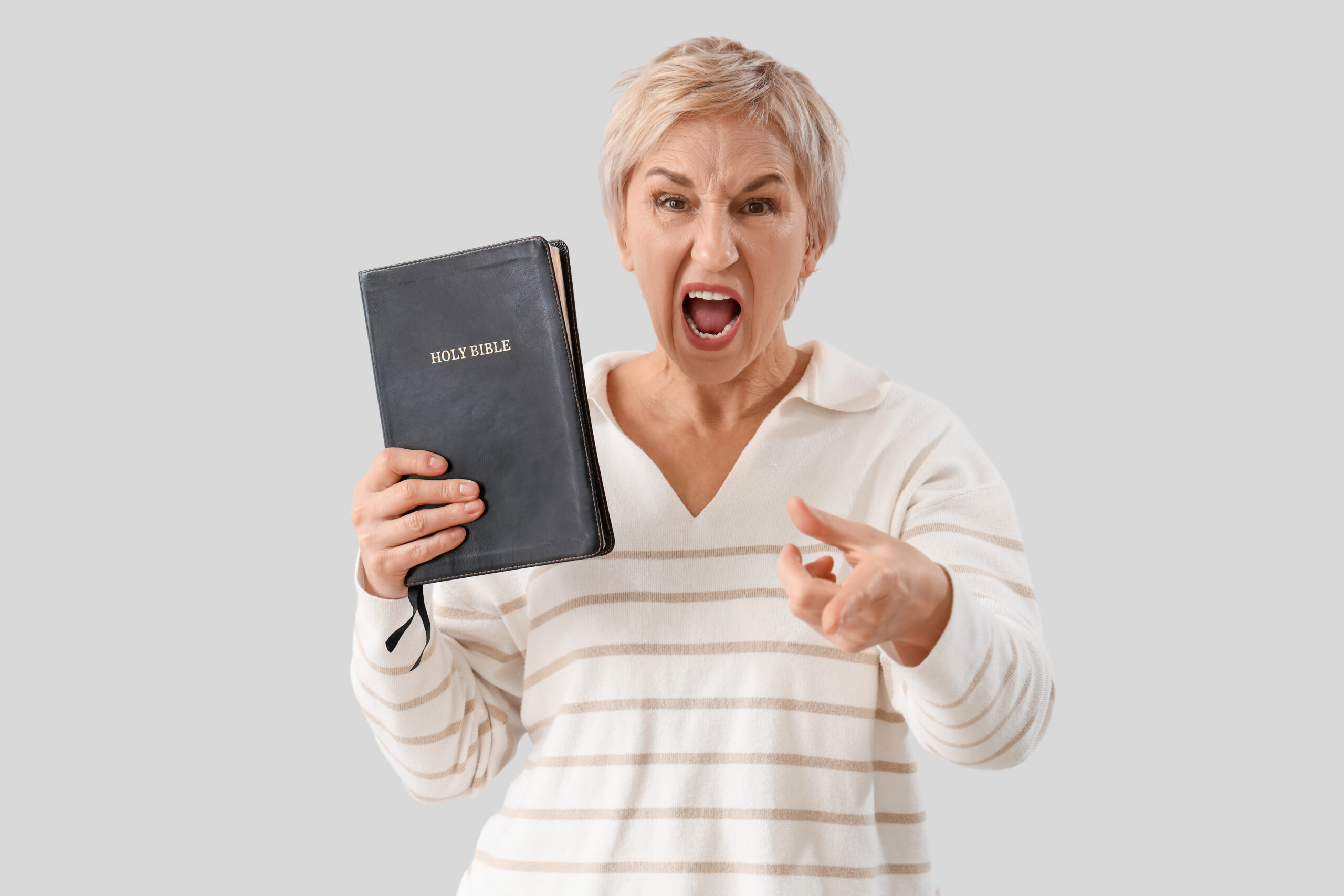 Angry mature woman with Holy Bible pointing at viewer on light background. Accusation concept